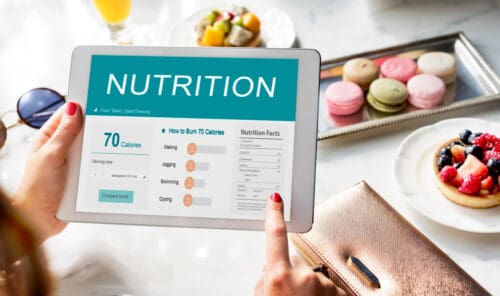 Persona consultando información nutricional en una tablet, mostrando datos sobre calorías y consumo energético, con postres y frutas frescas en la mesa